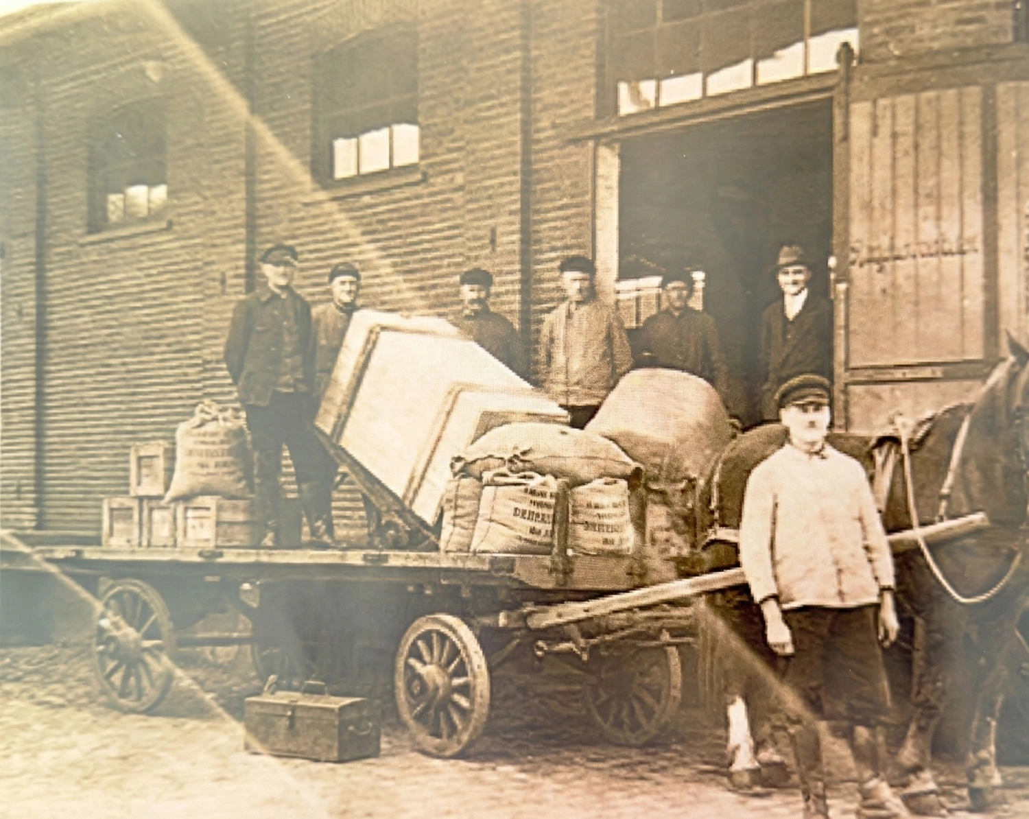 Progreen GmbH – Logistik – Dieses Bild zeigt eine Szene aus der Industrielogistik des frühen 20. Jahrhunderts: Arbeiter bereiten einen Pferdewagen für den Transport vor.